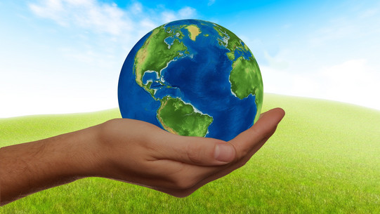 One hand holds a globe against the background of a green meadow with a blue sky.