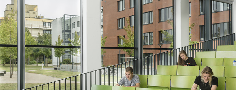 Drei Studierende sitzen in einem grün bestuhlten Hörsaal.