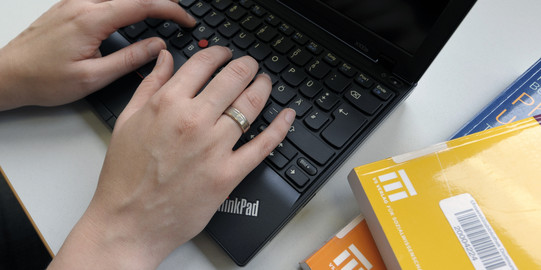 Eine Studentin tippt an einem Laptop und gelbe Bücher legen daneben.