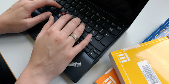 Eine Studentin tippt an einem Laptop und gelbe Bücher legen daneben.
