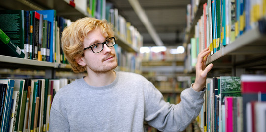 Bücherrecherche in der Bibliothek Studierender steht suchend an einem Bücherregal in der Bibliothek.